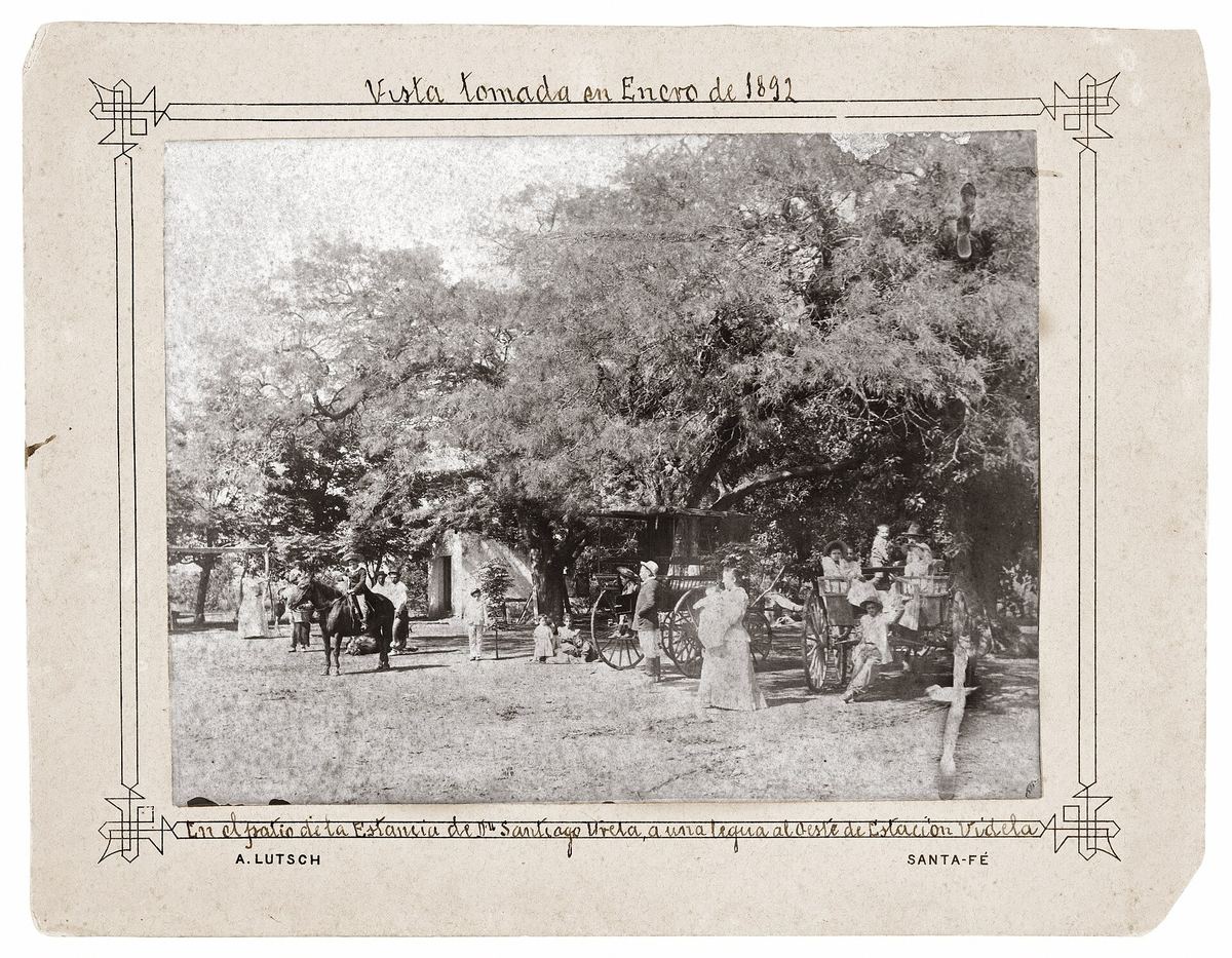 Fotografía antigua en blanco y negro de una estancia con árboles, carruajes y personas, con marco dibujado y leyenda manuscrita