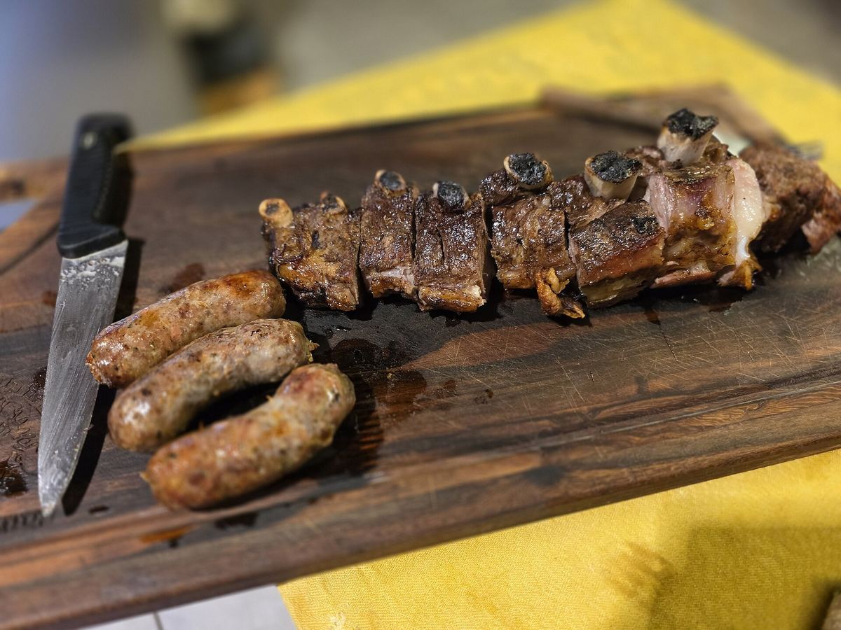 Tabla de madera con costillas de asado cortadas y chorizos a la parrilla, junto a un cuchillo