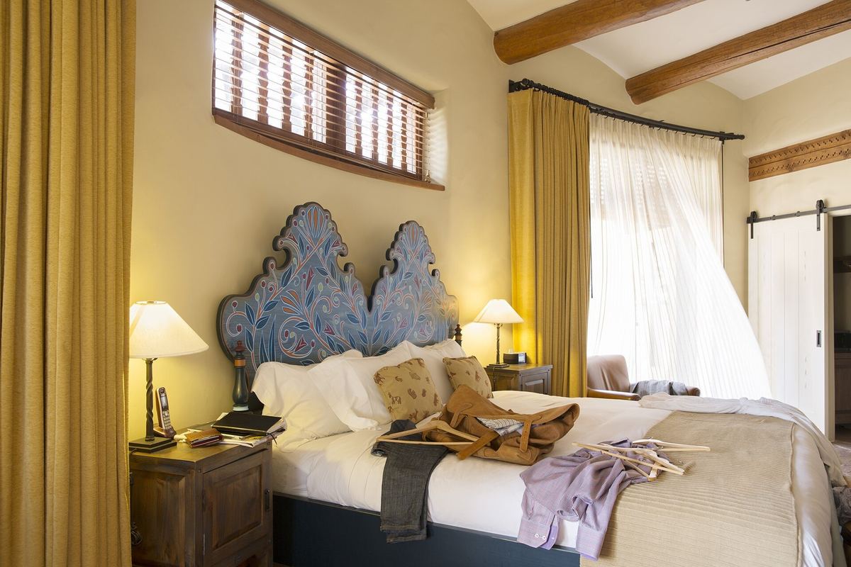 Habitación con cabecera tallada y pintada, vigas de madera a la vista y cortinas amarillas junto a la ventana